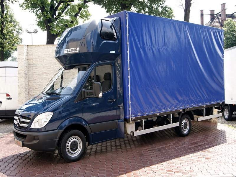 Niebieski samochód dostawczy Mercedes-Benz z plandeką, zaparkowany na brukowanej powierzchni, z logo 'LAMAR' nad przednią szybą.