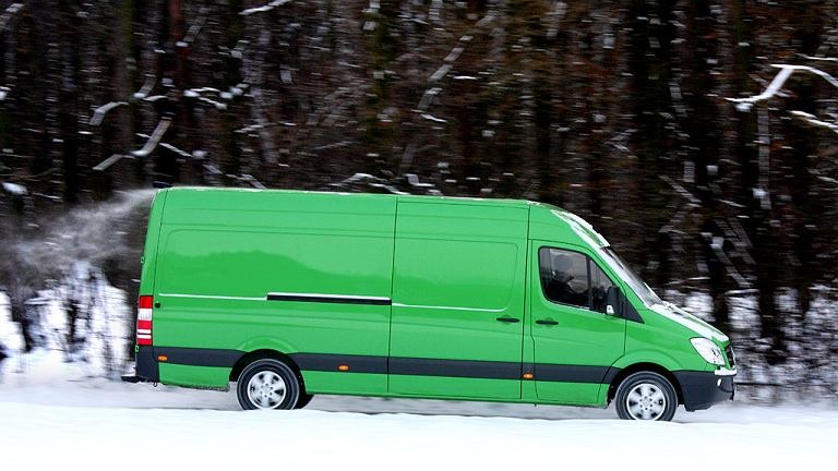 Zielony bus dostawczy Mercedes-Benz Sprinter jadący po ośnieżonej drodze na tle zimowego lasu.