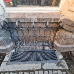 Metalowa krata okienna z dekoracyjnymi elementami spiralnymi, zabezpieczająca okno piwniczne w starym budynku z kamienną elewacją.