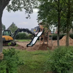 Żółta minikoparka przygotowuje teren pod plac zabaw z drewnianą konstrukcją ze zjeżdżalnią, widoczne kopce ziemi i drzewa.