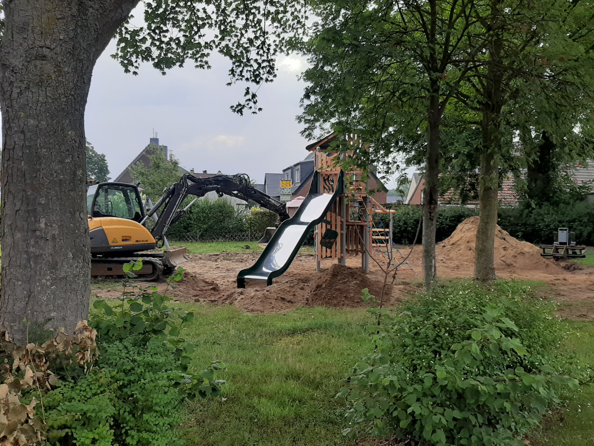 Żółta minikoparka przygotowuje teren pod plac zabaw z drewnianą konstrukcją ze zjeżdżalnią, widoczne kopce ziemi i drzewa.