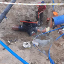 Wykop z widocznymi rurami kanalizacyjnymi z tworzywa sztucznego, złączkami, podsypką cementową i pracującym hydraulikiem z łopatą w tle, zabezpieczony taśmą ostrzegawczą.