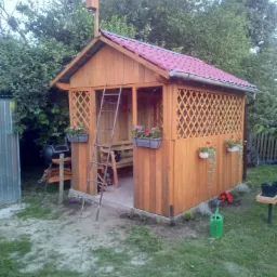 Drewniana altana ogrodowa z czerwoną dachówką i budką dla ptaków na szczycie, ozdobiona wiszącymi doniczkami z kwiatami, drabina oparta o ścianę, grill i ławka w tle.