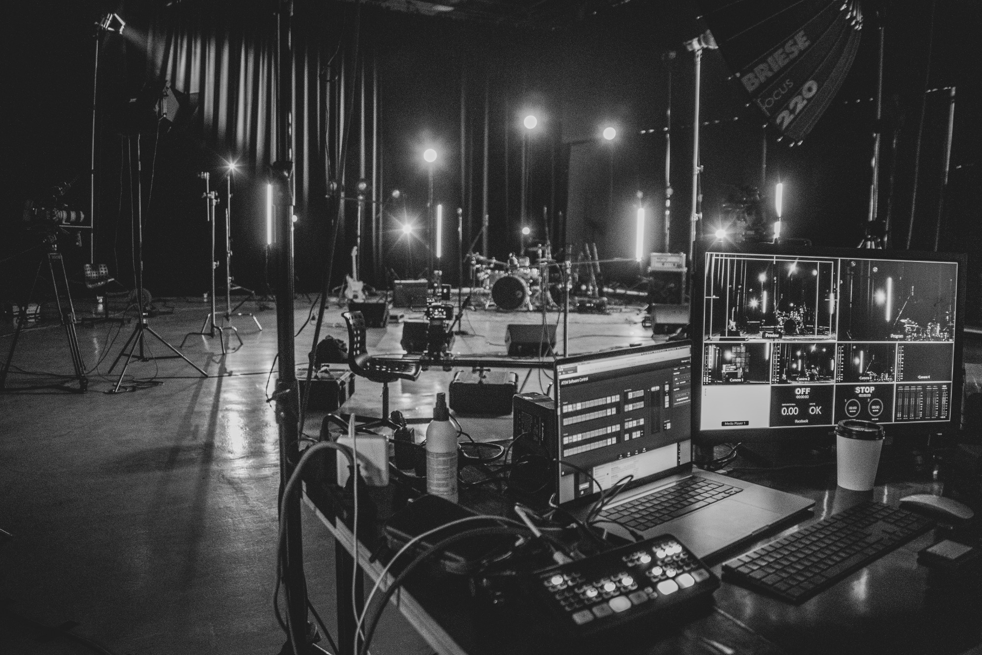 Czarno-białe zdjęcie zza kulis sesji nagraniowej zespołu muzycznego. Widoczny sprzęt oświetleniowy, monitory z podglądem z kamer, stanowisko realizatora dźwięku z laptopem i mikserem, oraz perkusja...