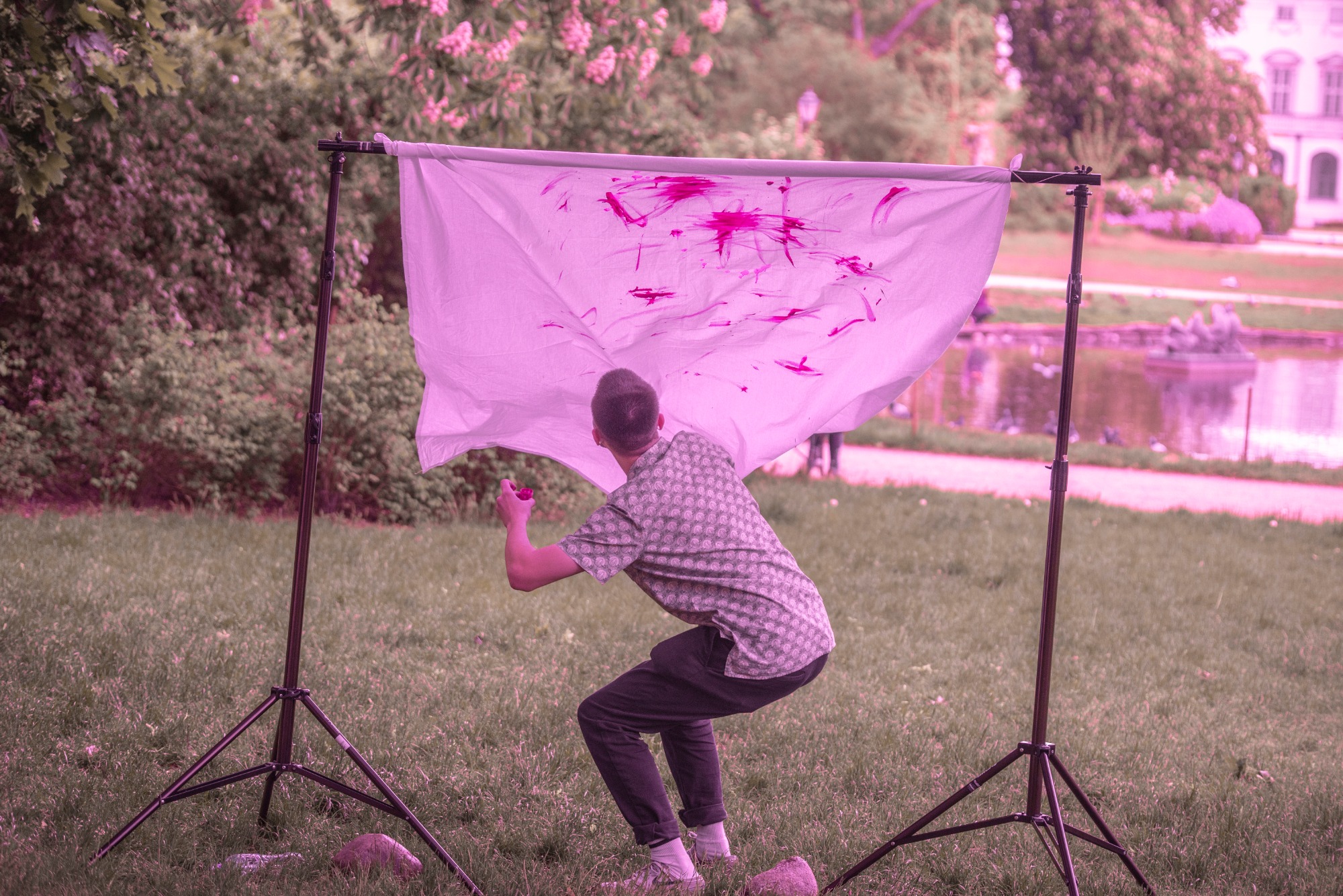 Młody mężczyzna w parku tworzy reportaż malarski, rozpryskując różową farbę na płótno zawieszone na statywach, z widocznym stawem i budynkiem w tle.
