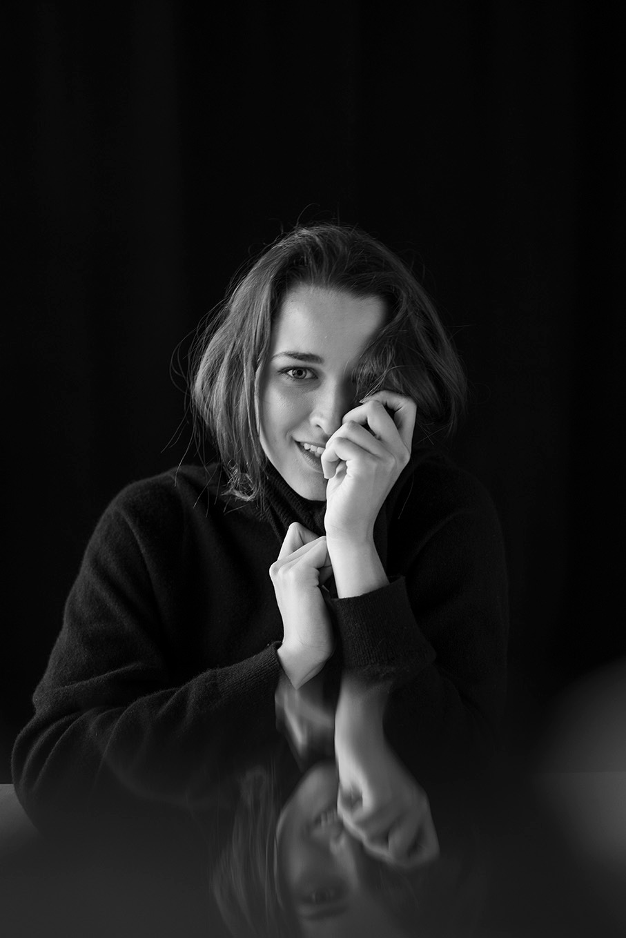 Czarno-biały portret uśmiechniętej młodej kobiety w czarnym golfie, z rękami przy twarzy i odbiciem w błyszczącej powierzchni.