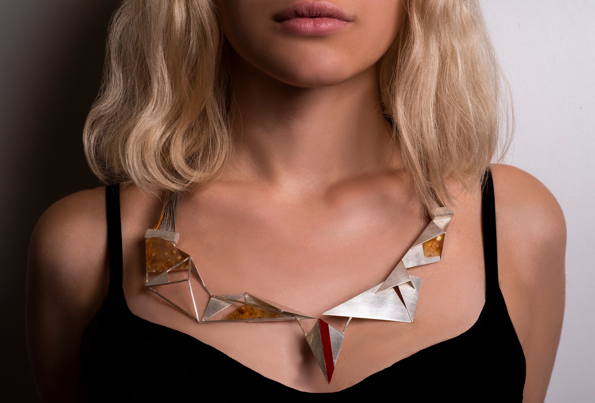 Kobieta z blond włosami nosi geometryczny naszyjnik z elementami z żywicy i czerwonym akcentem, na białym tle.