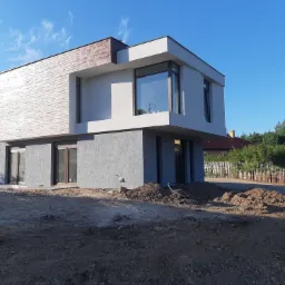 Dwukondygnacyjny dom o nowoczesnej architekturze z płaskim dachem, górna część wykończona teksturą przypominającą cegłę, dolna część w szarym tynku strukturalnym, duże okna, teren wokół domu...