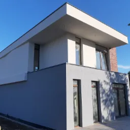 Dwukondygnacyjny dom o nowoczesnej architekturze z płaskim dachem, elewacja w odcieniach szarości i bieli, fragment ściany wykończony czerwoną cegłą, okna z ciemnymi ramami, błękitne niebo w tle.