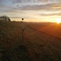 Profesjonalny odbiornik GNSS na tyczce geodezyjnej wbity w porośnięty trawą pagórek, w tle wschodzące słońce i lekka mgła nad łąką.