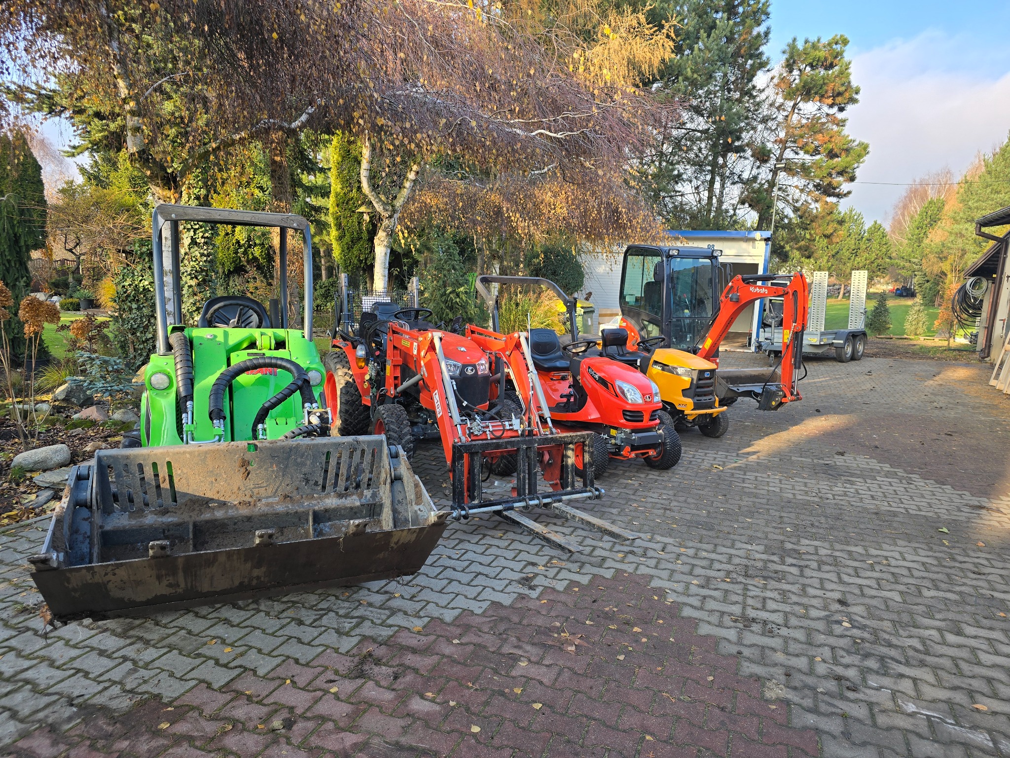 Park maszyn ogrodniczych: zielony miniładowarka, pomarańczowe traktory z osprzętem i żółta mini koparka na tle jesiennego ogrodu.