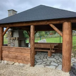Drewniana altana z grillem murowanym i ławkami, wykończona w rustykalnym stylu. Widoczne okrągłe słupy i kamienna podłoga.