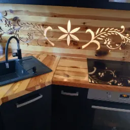 Wyjątkowa drewniana ściana nad blatem kuchennym z wyciętymi, podświetlonymi wzorami roślinnymi. Czarny zlew i płyta indukcyjna wkomponowane w blat.