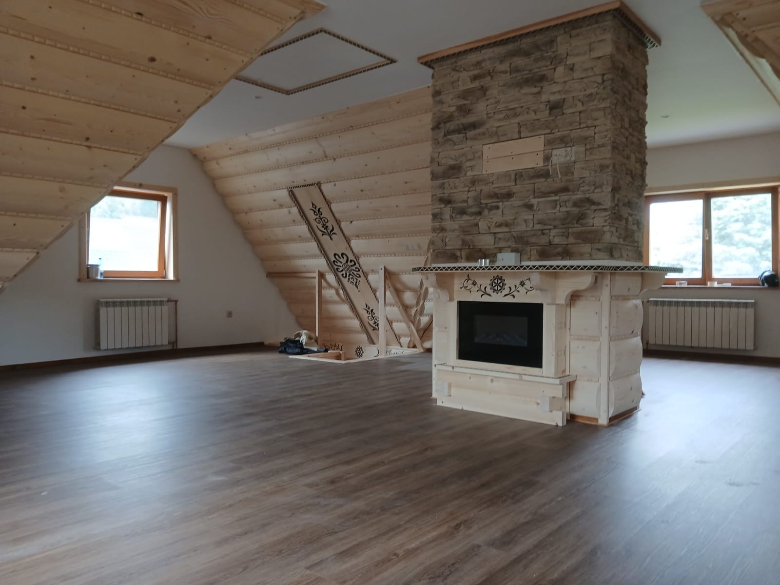 Wykończone poddasze w stylu góralskim: drewniane ściany, dekoracyjne schody, kominek obłożony kamieniem. Widok na okno i grzejnik. Parkiet na podłodze.