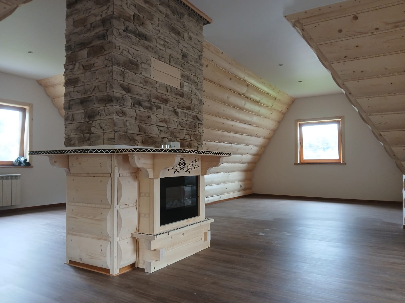 Drewniane wykończenie poddasza z kominkiem obłożonym kamieniem. Ściany i sufit z boazerii, podłoga z paneli. Widoczne okno i fragment grzejnika.