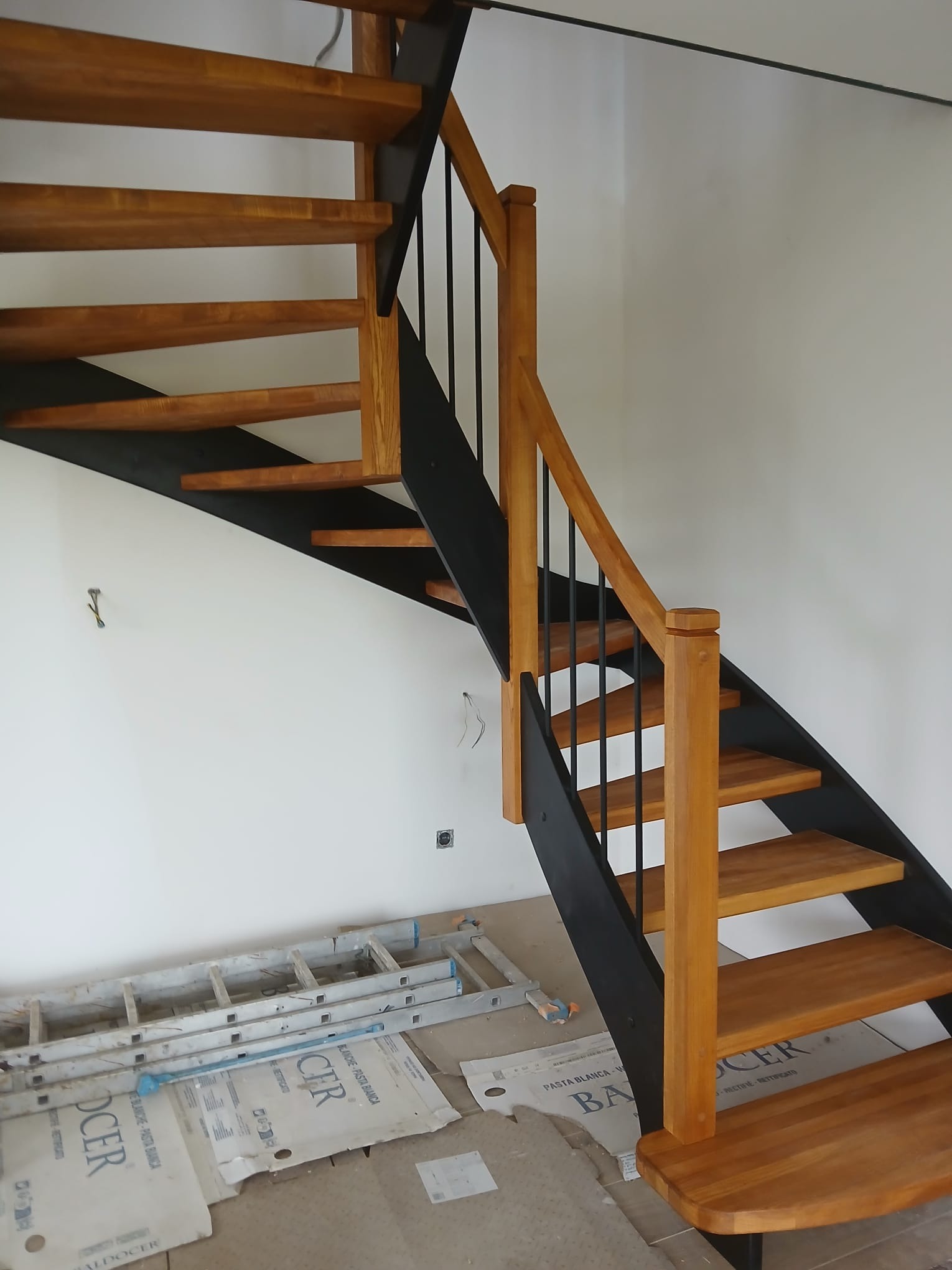 Schody drewniane z czarną konstrukcją i jasnymi stopniami, widoczne z boku, z elementami wykończeniowymi w tle. Układ schodów spiralny.