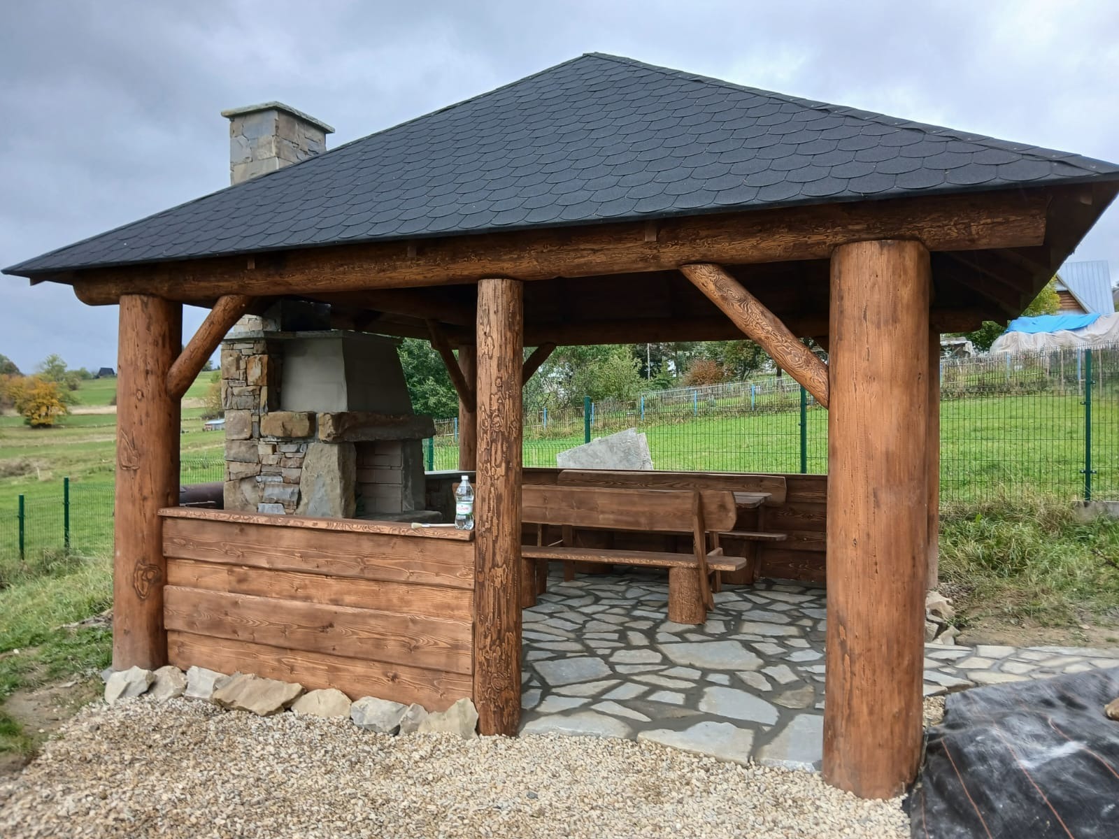 Drewniana altana z grillem murowanym i ławkami, wykończona w rustykalnym stylu. Widoczne okrągłe słupy i kamienna podłoga.