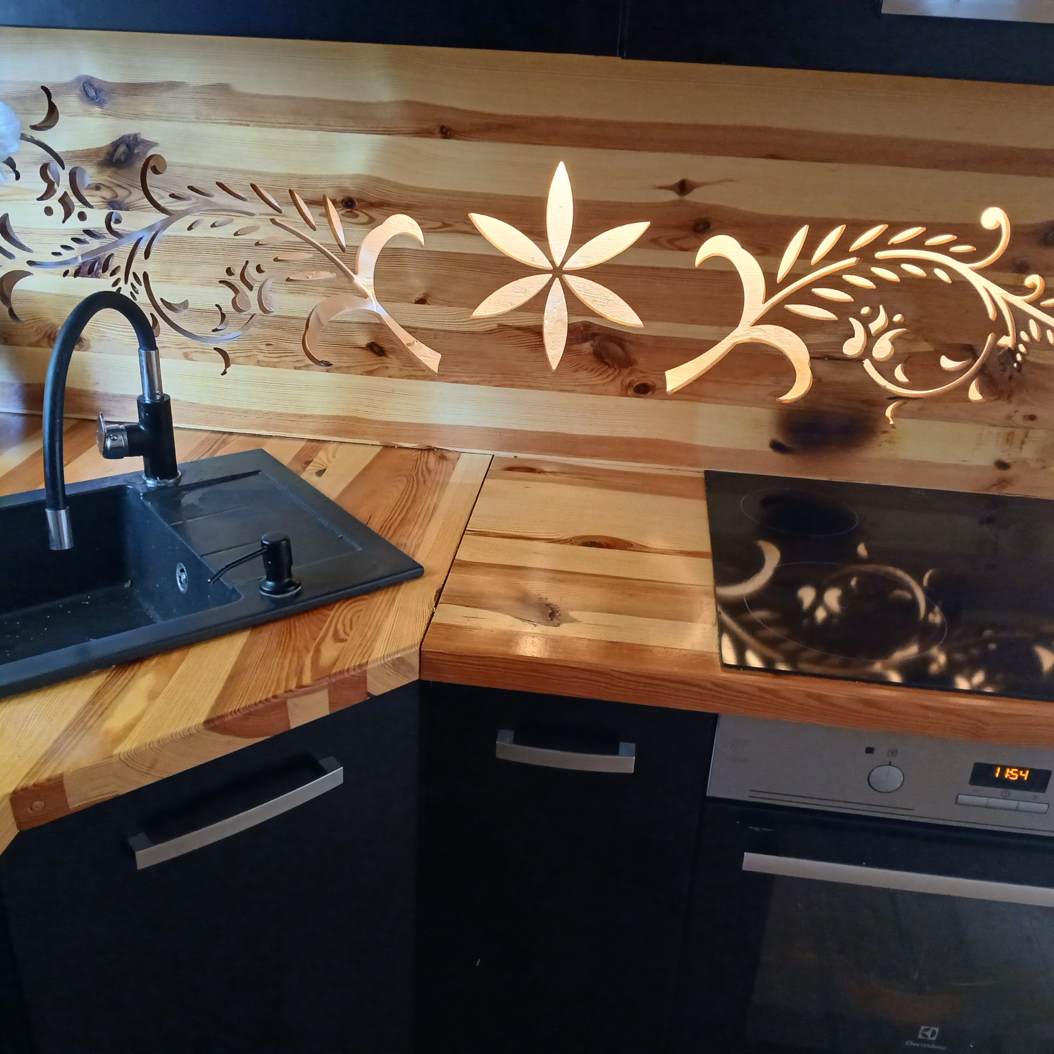Wyjątkowa drewniana ściana nad blatem kuchennym z wyciętymi, podświetlonymi wzorami roślinnymi. Czarny zlew i płyta indukcyjna wkomponowane w blat.