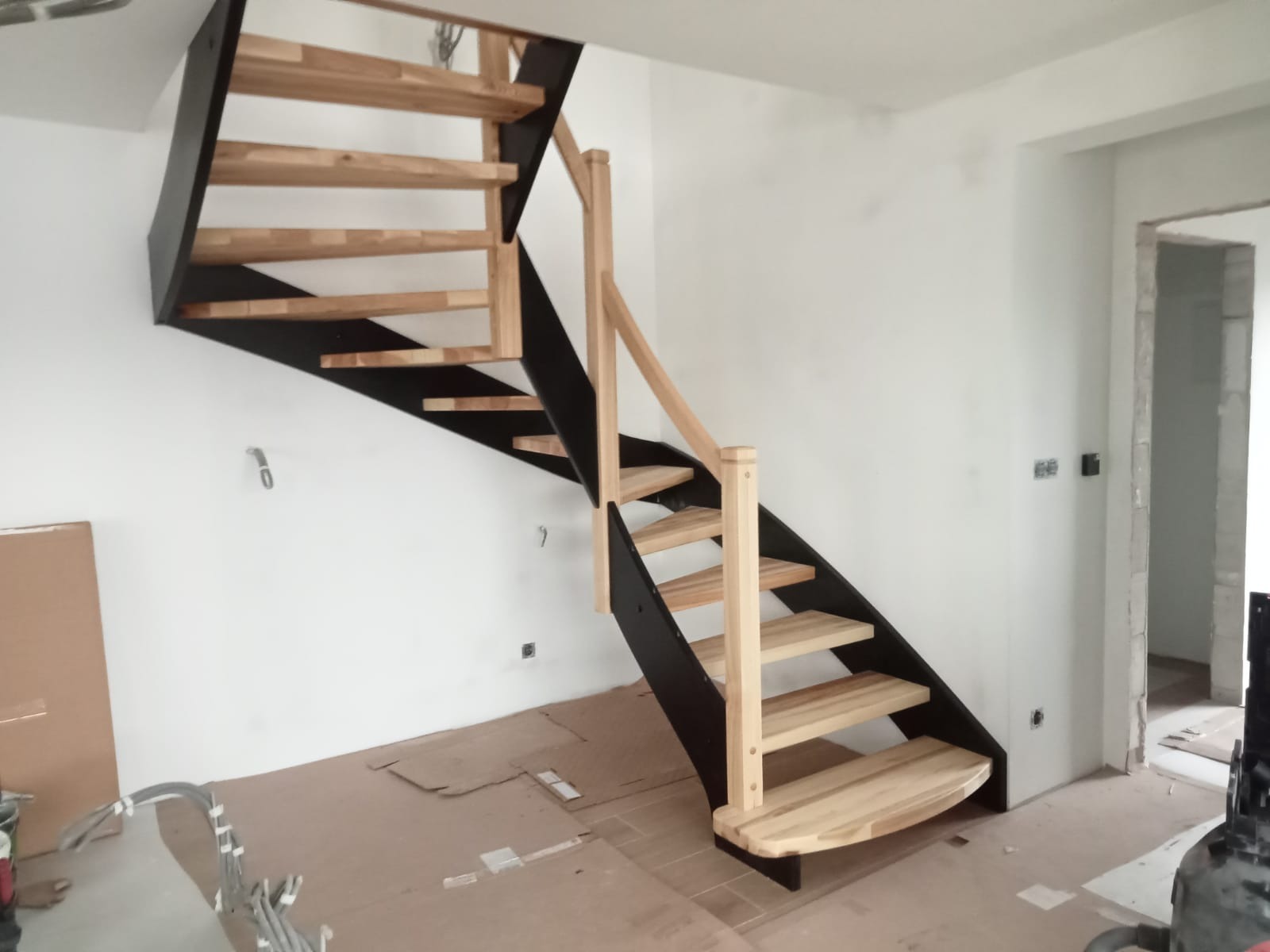 Nowoczesne schody z drewnianymi stopniami i czarną metalową konstrukcją, zamontowane w jasnym wnętrzu domu w trakcie wykończenia.