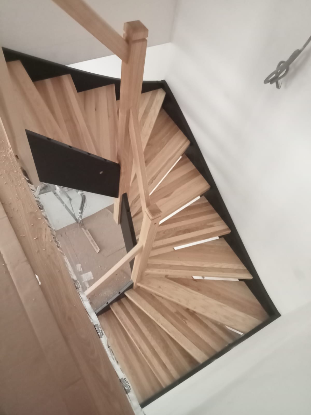 Widok z góry na jasne, drewniane schody spiralne z czarnymi elementami, umieszczone w jasnym wnętrzu. Nietypowe ujęcie podkreśla geometrię konstrukcji.