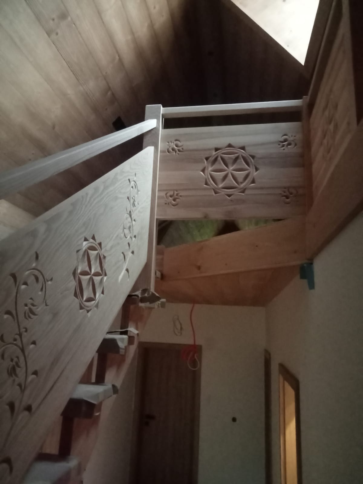 Drewniane schody z dekoracyjnymi, rzeźbionymi balustradami i motywem kwiatowym, widziane z dołu, w jasnym wnętrzu z drewnianym sufitem.