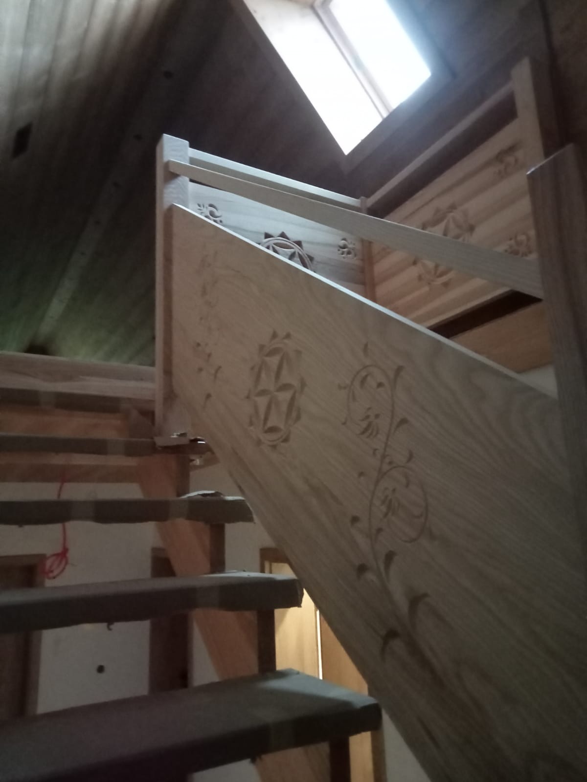 Drewniane schody z dekoracyjnymi, rzeźbionymi balustradami, oświetlone przez okno dachowe. Widoczny fragment drewnianego sufitu i konstrukcji schodów.