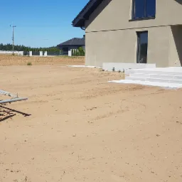 Przygotowanie terenu pod trawnik: mały pomarańczowy traktor Kubota z broną wirnikową spulchnia ziemię przed nowym domem z betonowymi schodami i tarasem.