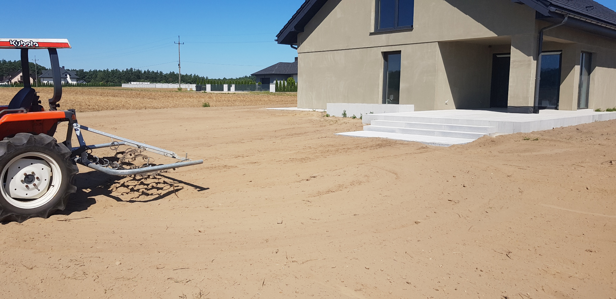 Przygotowanie terenu pod trawnik: mały pomarańczowy traktor Kubota z broną wirnikową spulchnia ziemię przed nowym domem z betonowymi schodami i tarasem.