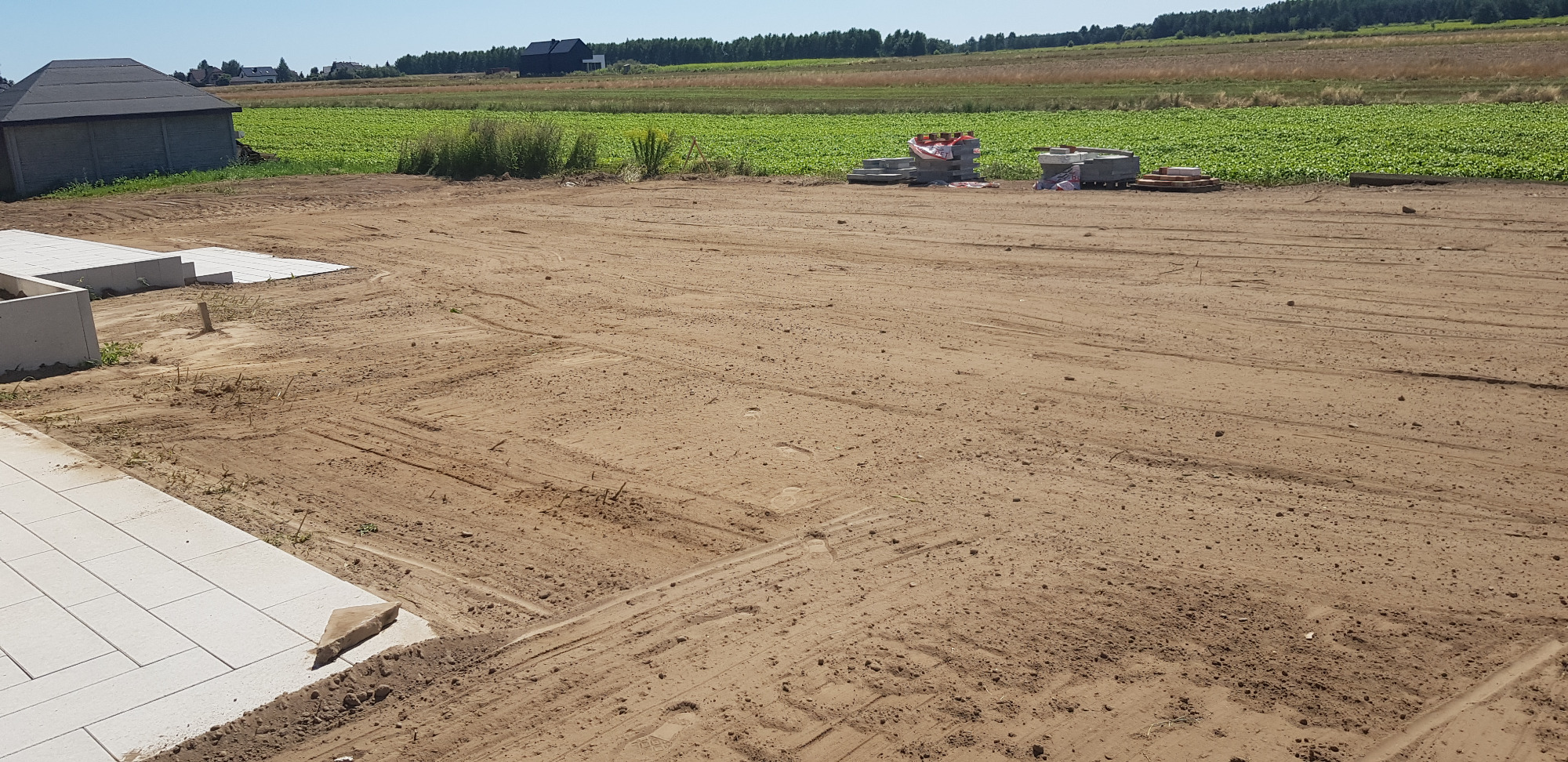 Przygotowany teren pod budowę z ułożonymi płytami chodnikowymi i paletami z materiałami budowlanymi w tle, widok na pole uprawne.