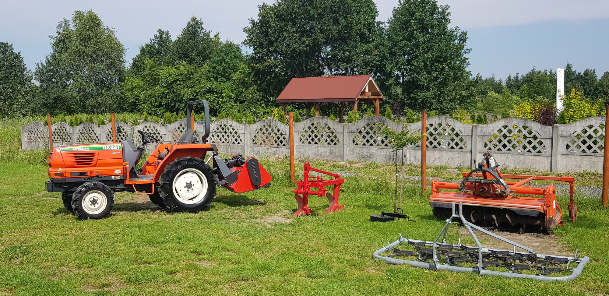 Pomarańczowy traktor Hitachi NX221 z osprzętem rolniczym na trawniku, w tle betonowy płot i altana z czerwonym dachem.