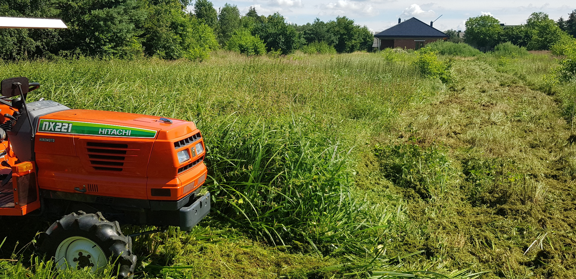 Pomarańczowy traktor Hitachi NX221 na zarośniętej łące, z widocznym śladem po koszeniu i domem w tle.
