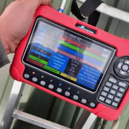 Miernik sygnału satelitarnego DVB-S2 w czerwonej obudowie, trzymany w dłoni na tle drabiny i zielonego dachu, wyświetlający parametry sygnału z satelity Ku Hotbird 13.