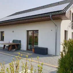 Biały dom jednorodzinny z panelami fotowoltaicznymi na dachu, widok z zewnątrz, stół ogrodowy z krzesłami na kółkach przy ścianie, oraz antena telewizyjna na bocznej ścianie domu.