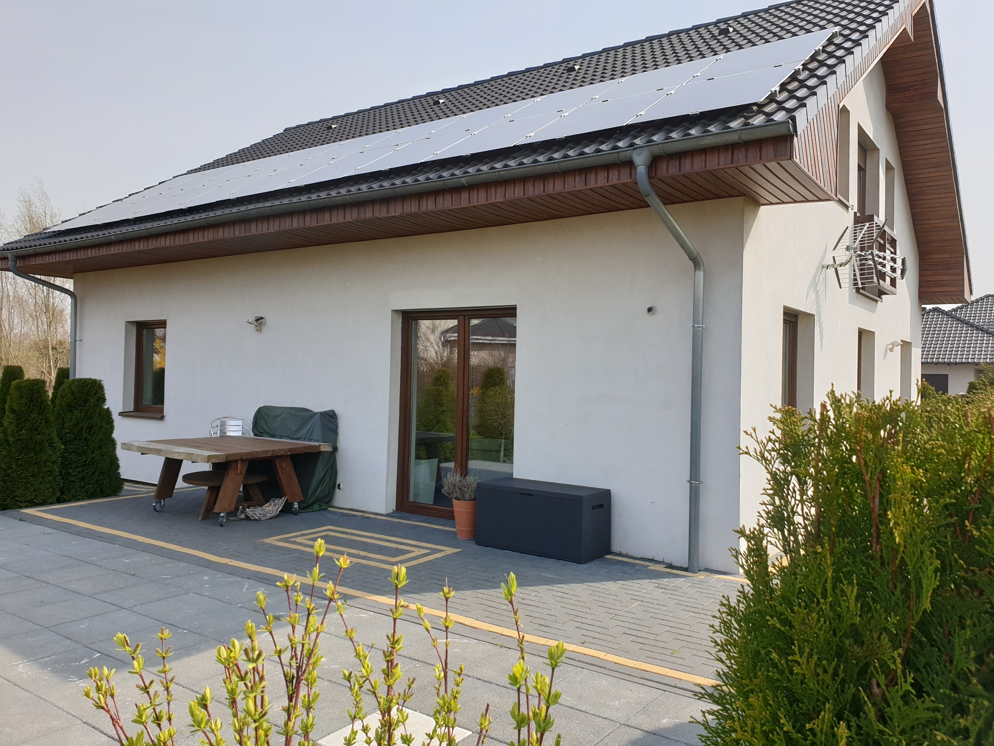 Biały dom jednorodzinny z panelami fotowoltaicznymi na dachu, widok z zewnątrz, stół ogrodowy z krzesłami na kółkach przy ścianie, oraz antena telewizyjna na bocznej ścianie domu.