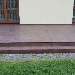 Brązowe płytki ceramiczne na schodach i tarasie przed domem, widok z poziomu trawnika, zarys okien z brązowymi ramami w tle.