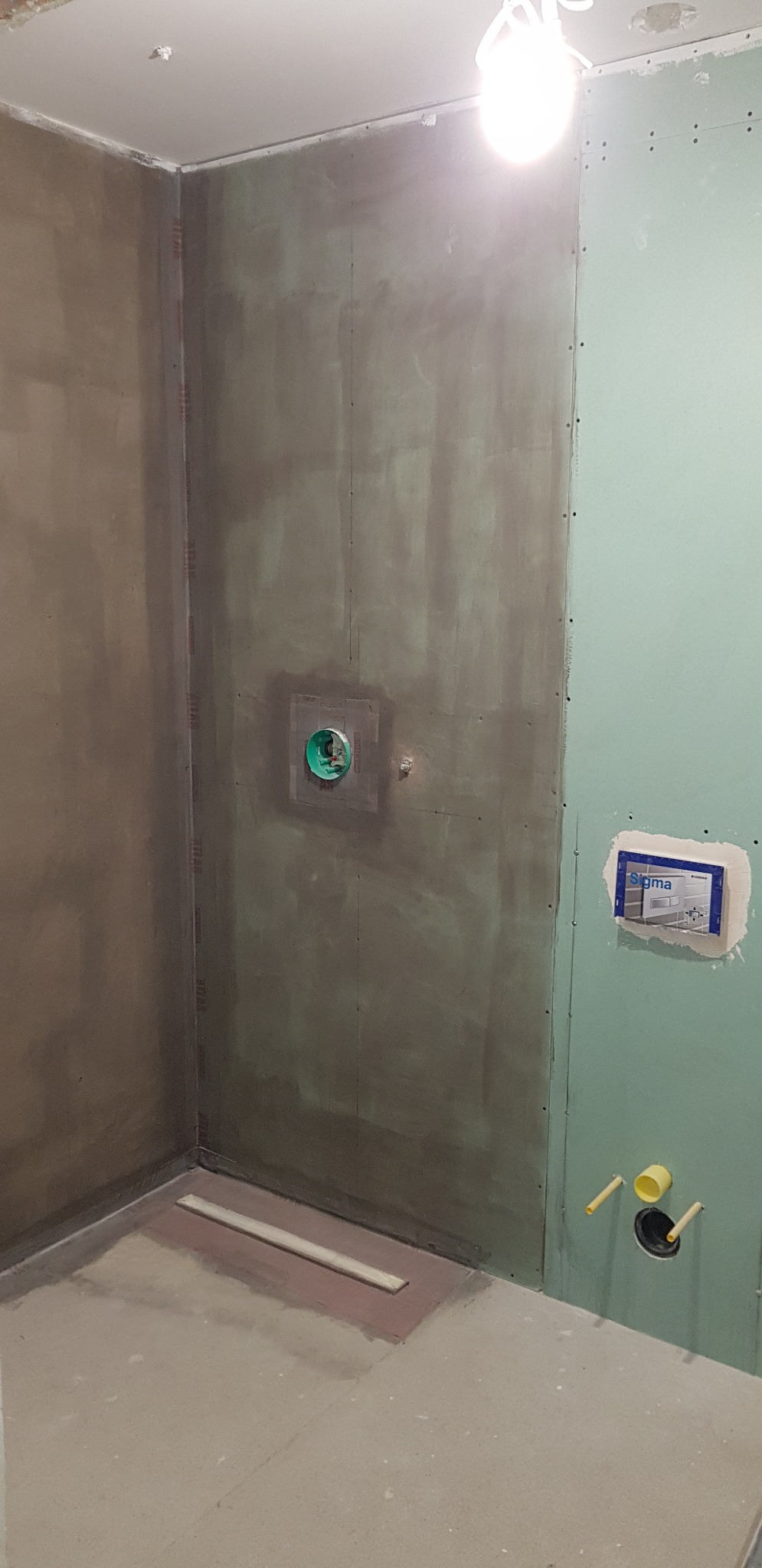 Surowy stan łazienki w trakcie remontu: ściany pokryte szarym tynkiem i zielonymi płytami gipsowo-kartonowymi, widoczne instalacje wodno-kanalizacyjne i odpływ liniowy w podłodze.