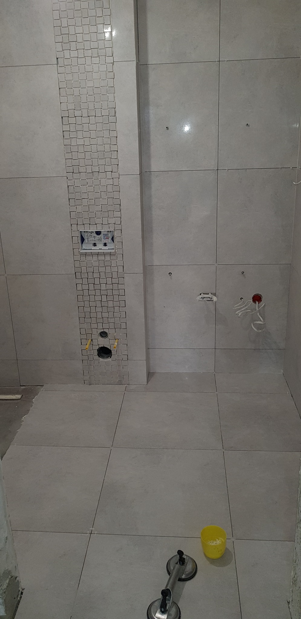 Ściana łazienki w trakcie układania płytek ceramicznych w odcieniach szarości, z dekoracyjnym pasem mozaiki i widocznymi otworami na instalacje. Na podłodze przyssawka do przenoszenia płytek...