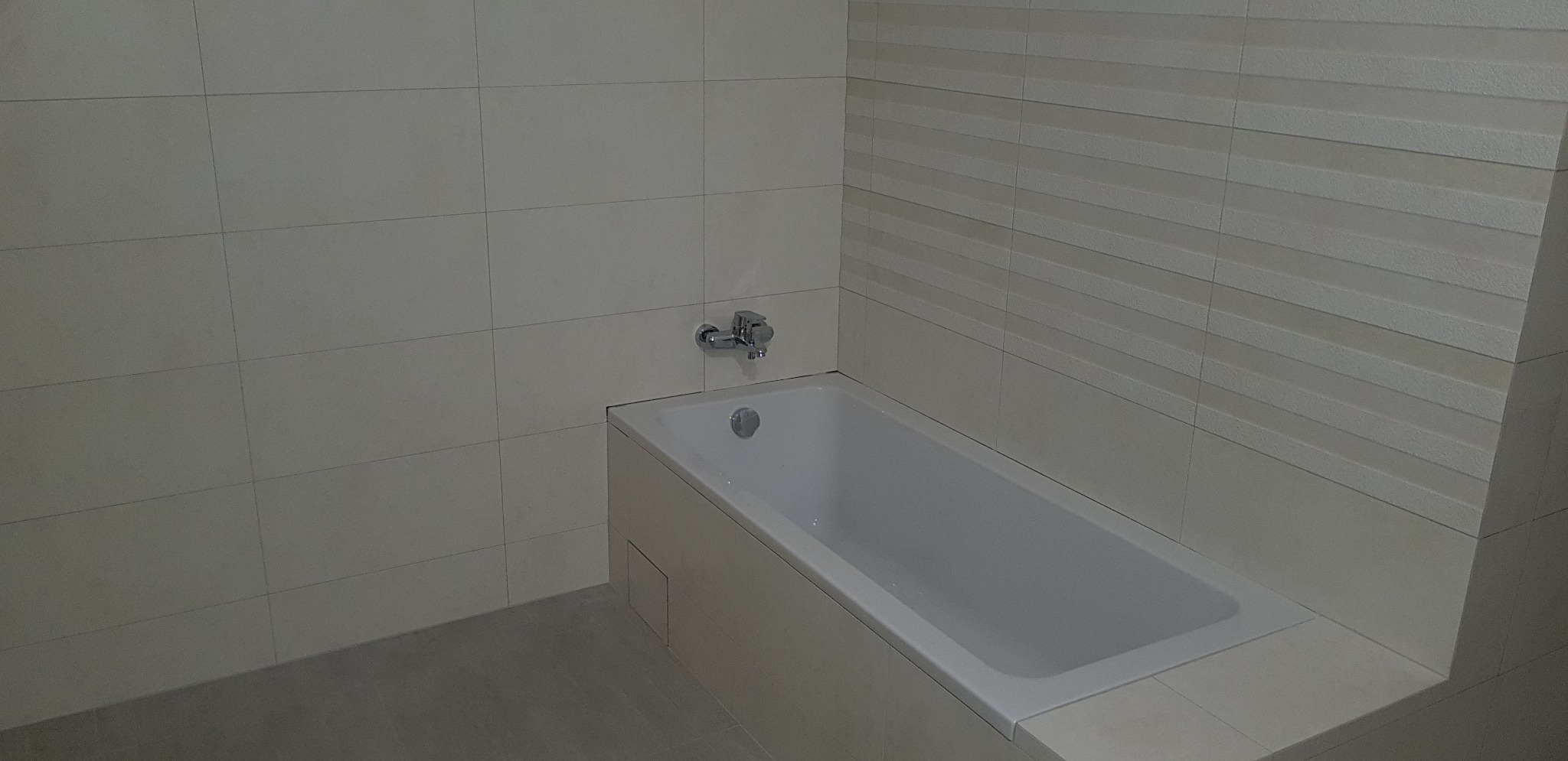 Nowoczesna, minimalistyczna łazienka z wanną obudowaną płytkami ceramicznymi, ściany wyłożone jasnymi płytkami o zróżnicowanej fakturze, chromowana bateria wannowa.