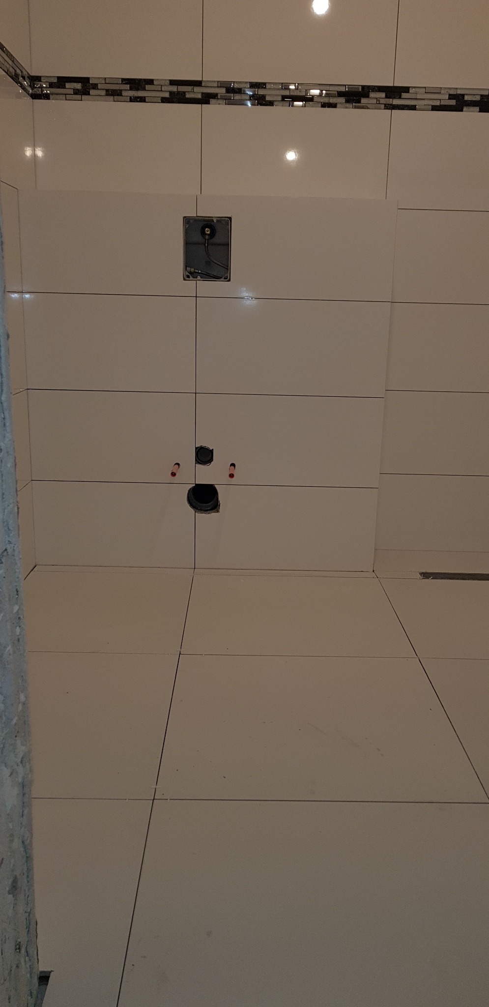 Białe płytki ceramiczne ułożone na ścianie i podłodze łazienki, widoczne otwory na instalacje wodno-kanalizacyjne i elektryczne, dekoracyjny pasek mozaiki na górze ściany.