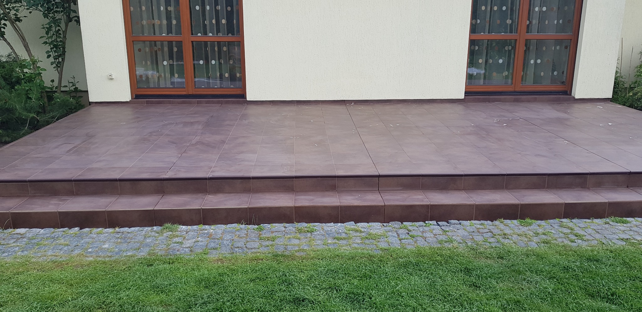 Brązowe płytki ceramiczne na schodach i tarasie przed domem, widok z poziomu trawnika, zarys okien z brązowymi ramami w tle.