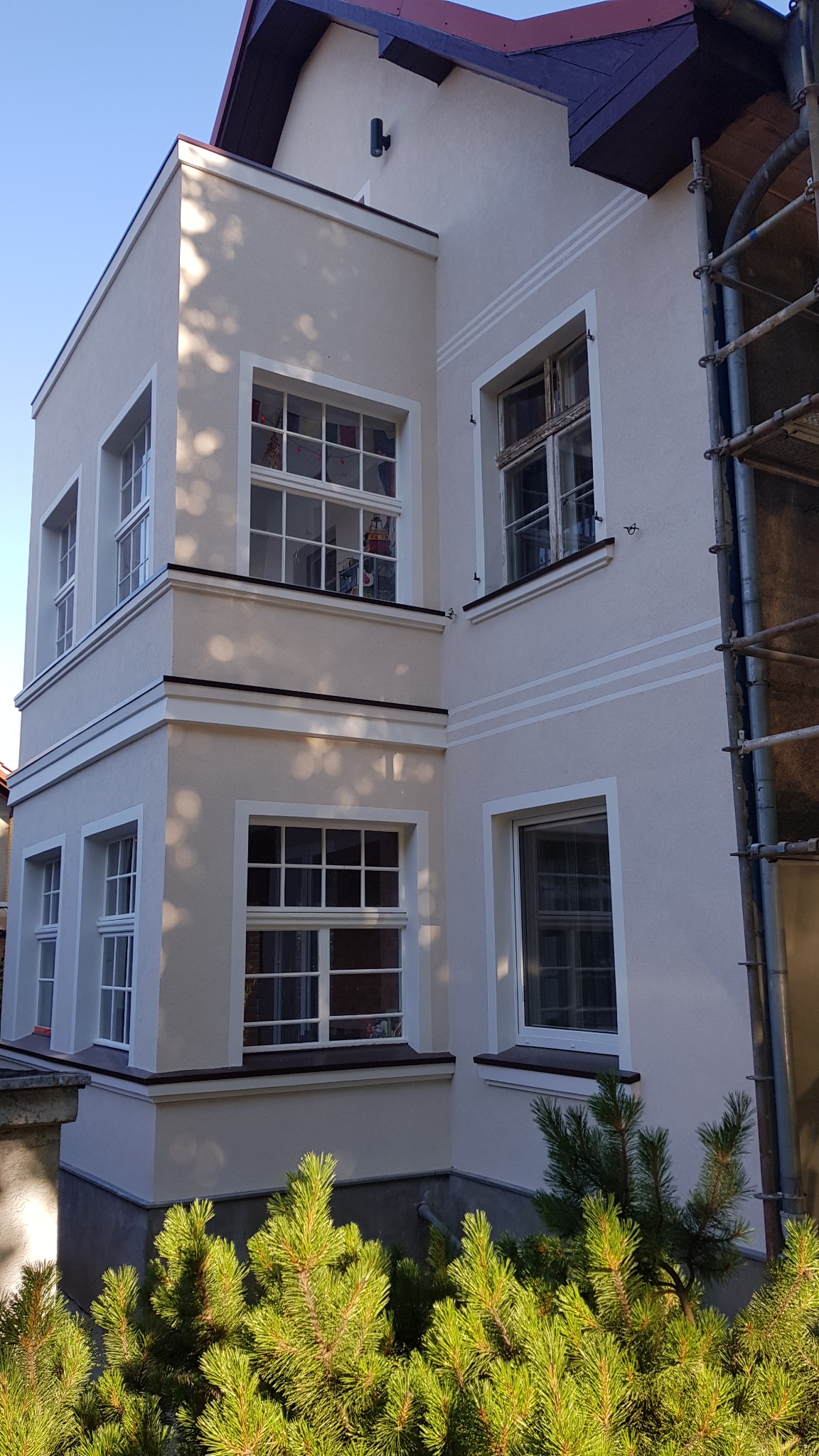 Odnowiona elewacja kamienicy w Szczecinie, z jasną, gładką powierzchnią, białymi obramowaniami okien i dekoracyjnymi poziomymi pasami, widoczne rusztowanie z prawej strony, na pierwszym planie...