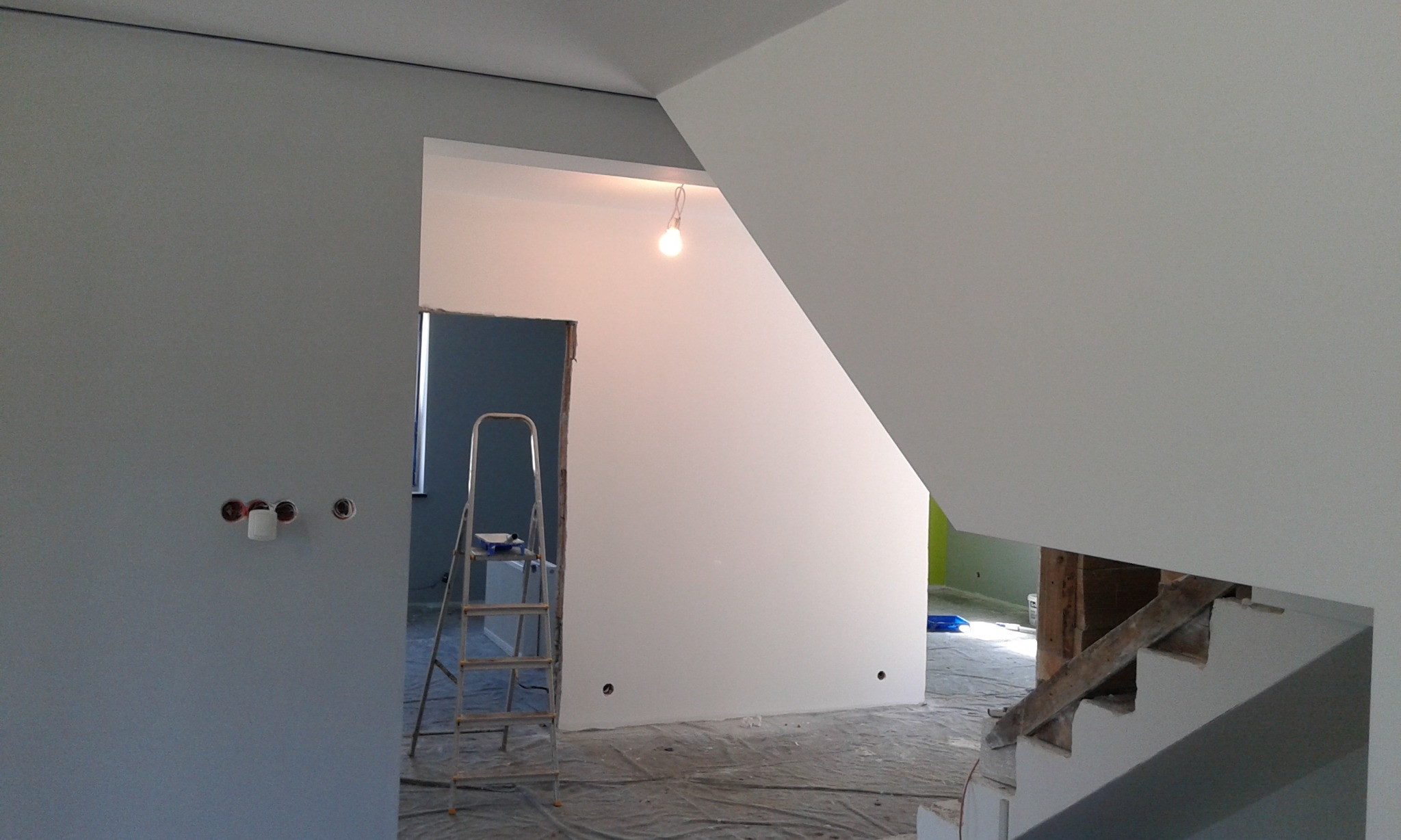 Wnętrze w trakcie malowania, z widocznymi gładkimi, świeżo pomalowanymi ścianami w odcieniach szarości i bieli, drabina malarska z kuwetą, zabezpieczona folią podłoga i surowe schody w tle.