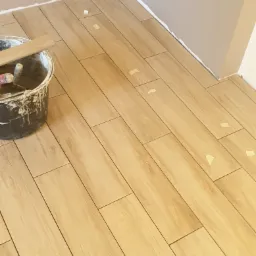 Układanie podłogi z płytek imitujących drewno w trakcie prac, widoczne taśmy zabezpieczające i wiadro z zaprawą.