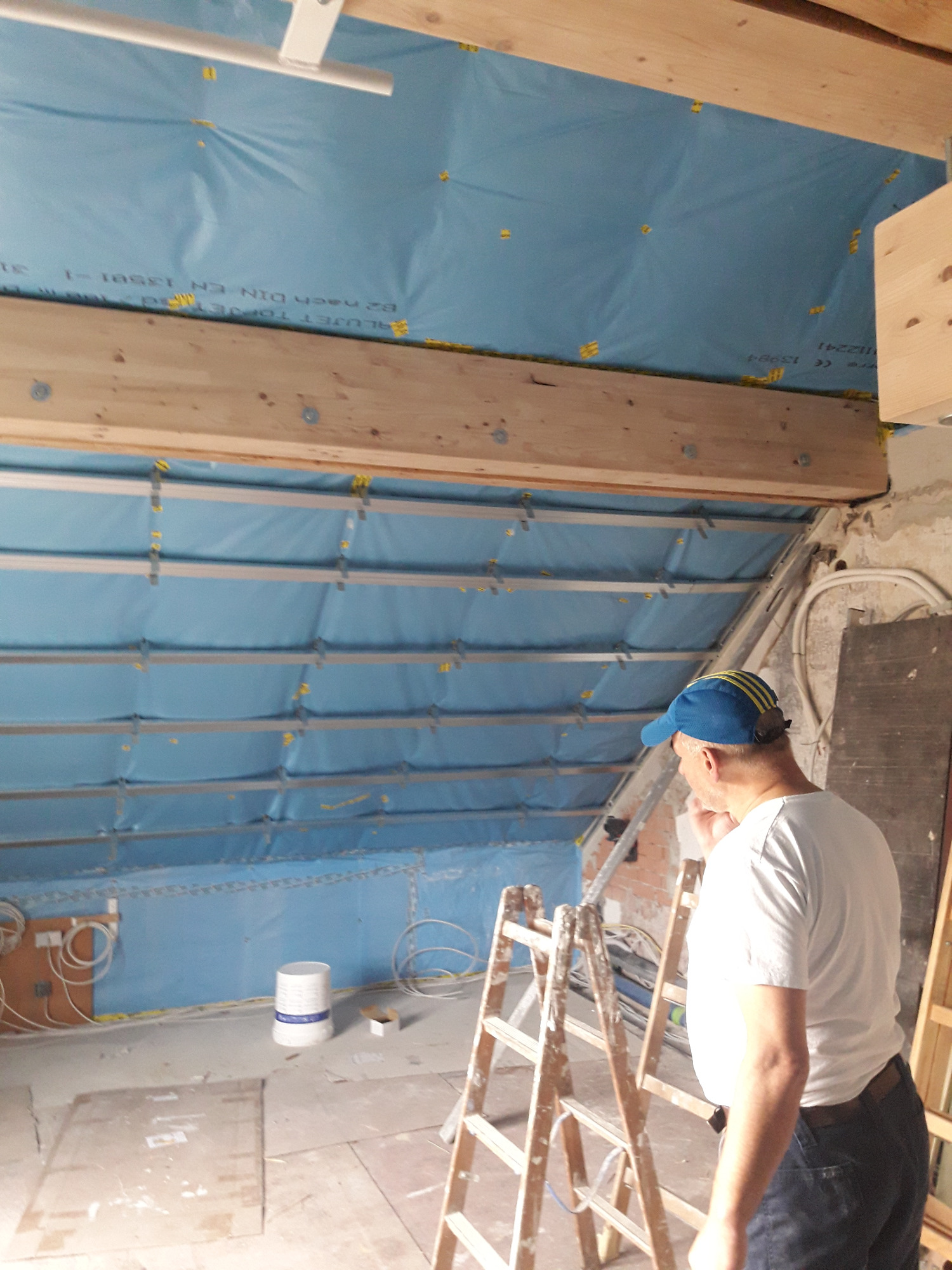 Montaż izolacji termicznej na poddaszu z widoczną konstrukcją drewnianą i aluminiowymi profilami, drabina oparta o ścianę.