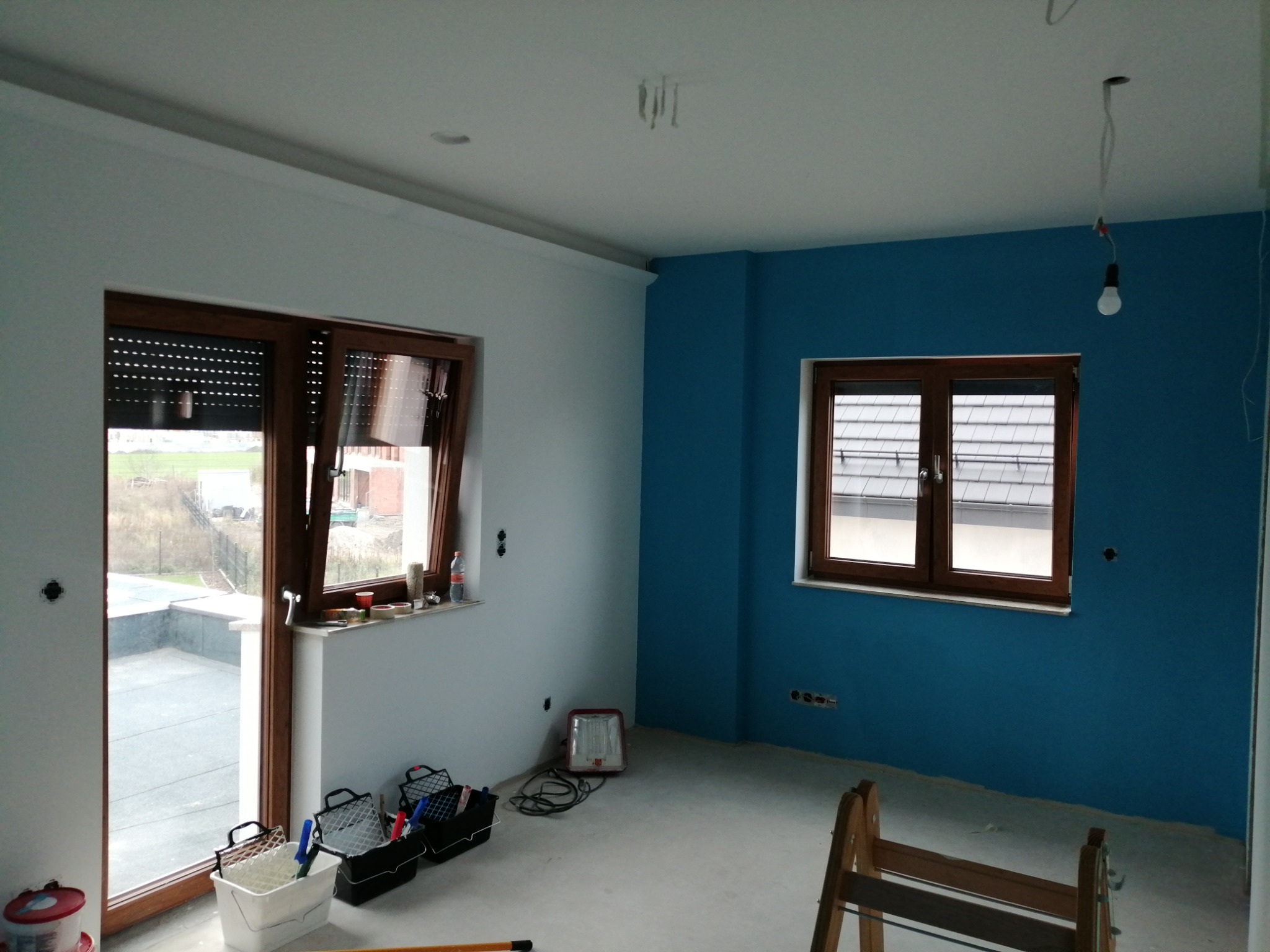 Pokój w trakcie odświeżania: jedna ściana pomalowana na intensywny niebieski kolor, druga biała, widoczne narzędzia malarskie, okna z drewnianymi ramami i roletami, surowa żarówka zwisająca z sufitu.
