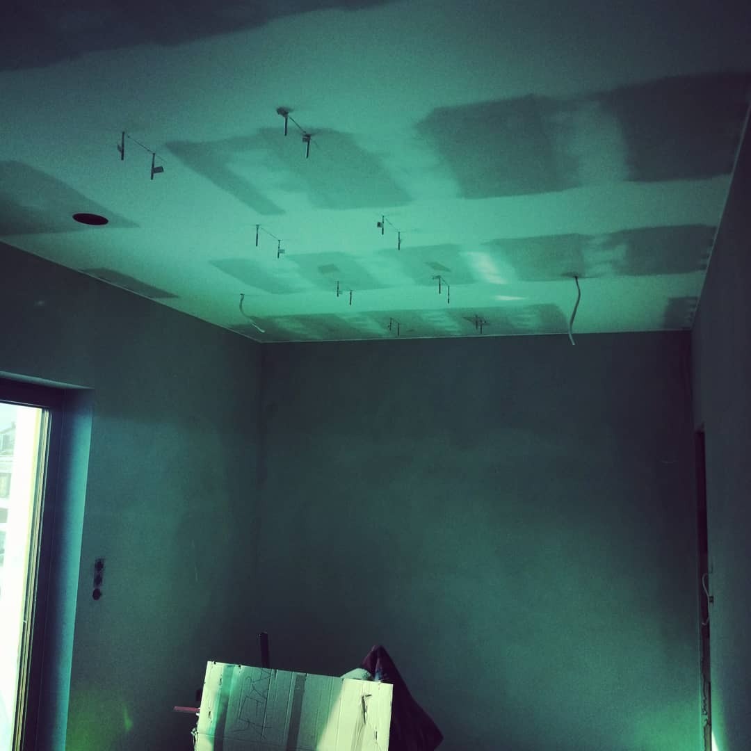 Pokój w trakcie prac przygotowawczych: surowa ściana, częściowo położona płyta na suficie, widoczne zawiesia mocujące, kabel zwisający z sufitu, oraz kartonowe zabezpieczenia na podłodze.