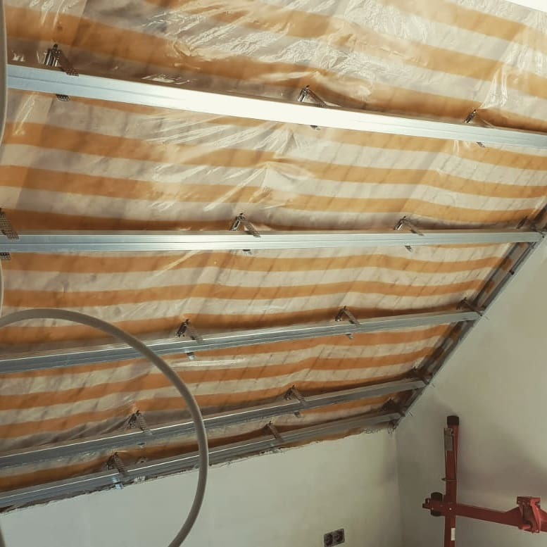 Montaż metalowego stelaża pod płytę karton gips na skośnym poddaszu, zabezpieczonego folią malarską w biało-pomarańczowe pasy.