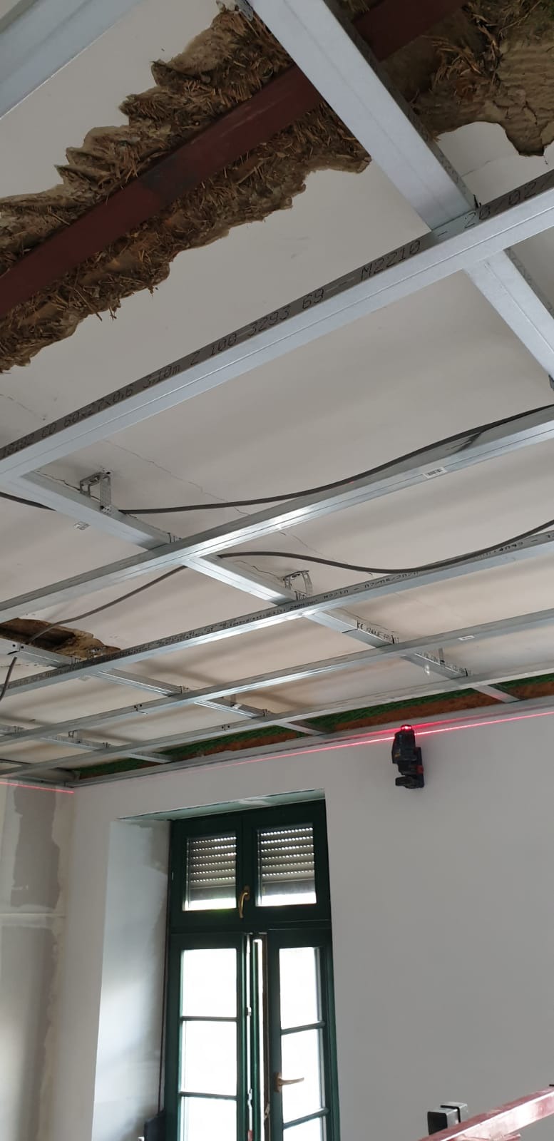 Montaż metalowej konstrukcji sufitu podwieszanego, widoczna izolacja i laserowy niwelator wyznaczający poziom na ścianie obok okna.