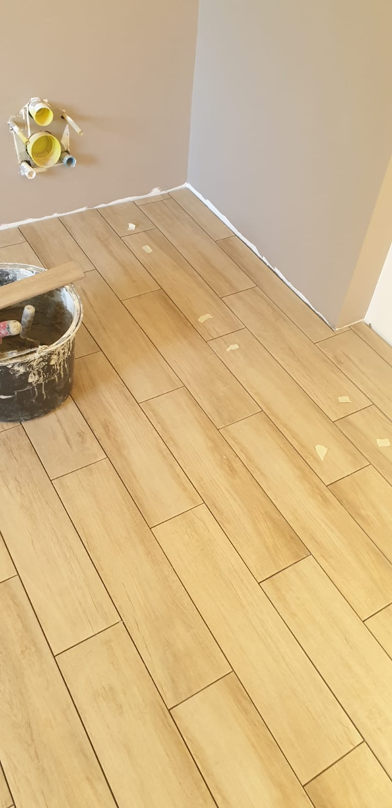 Układanie podłogi z płytek imitujących drewno w trakcie prac, widoczne taśmy zabezpieczające i wiadro z zaprawą.