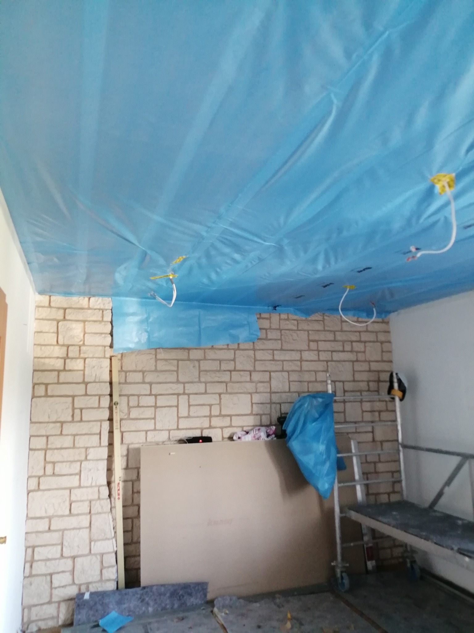 Przygotowanie do montażu płyty karton gips na suficie, zabezpieczonym niebieską folią, widoczna ściana z imitacji kamienia, płyta gipsowa oparta o ścianę oraz rusztowanie.